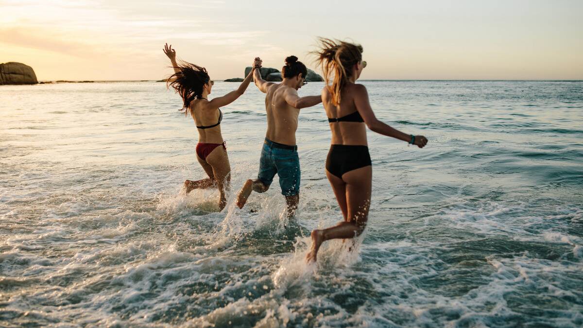 Three friends in bathing suits all holding hands as they run into the water.