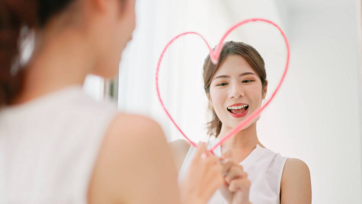 A woman smiling as she draws a heart around her face in a mirror with lipstick.