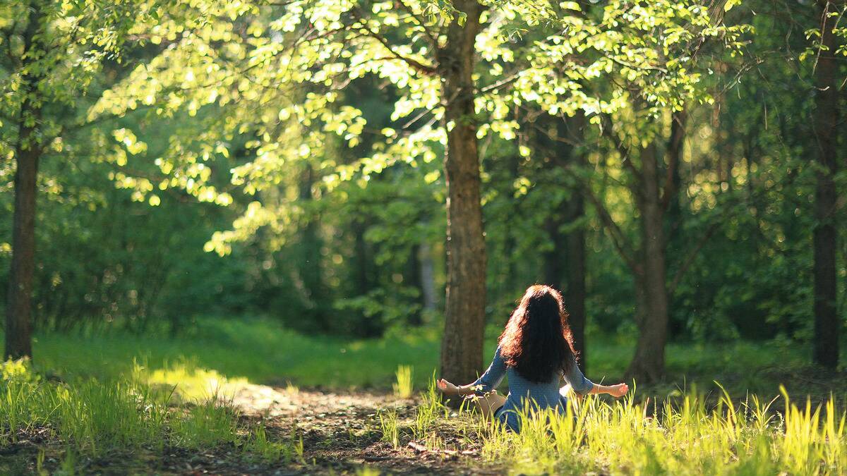 A woman sitting on the ground outside in a meditation pose, facing away from the camera.