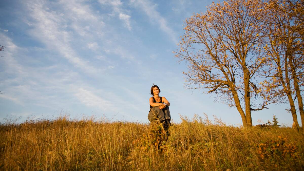 A woman sitting on a tree stump in the middle of a field of tall grass under a light blue sky.