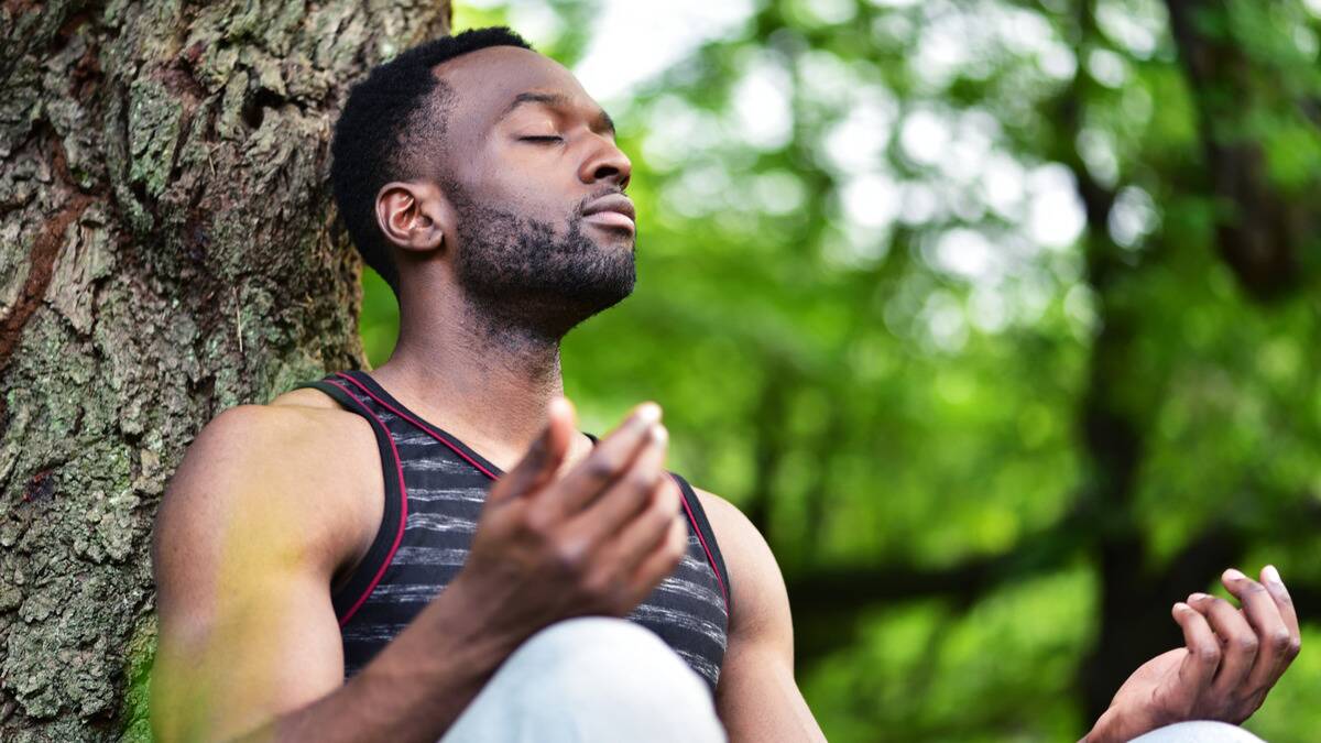 A low, close shot of a man sitting at the base of a tree, eyes closed, hand posed on his knee, meditating.