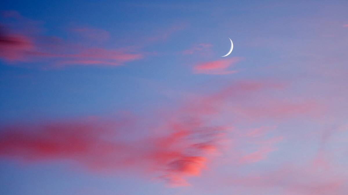 A thin sliver of a crescent moon high in a blue and pink sky.