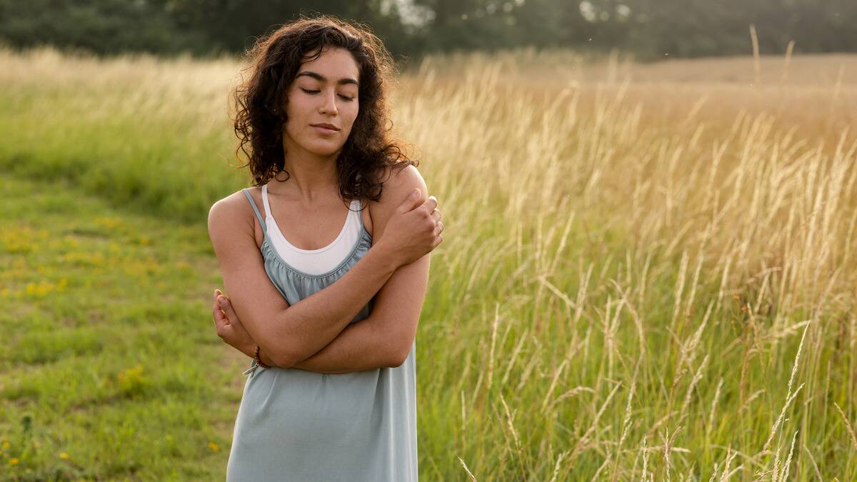 A woman standing in a field, smiling softly as she hugs herself.