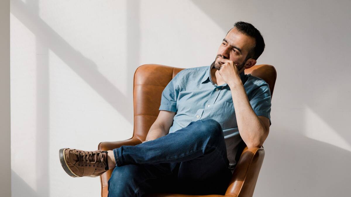 A man sitting in a chair by a large window, looking out it with a hand on his chin, clearly thinking.