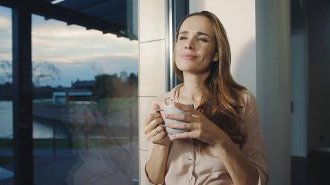 A woman standing inside, leaning against a wall by a large window, holding a mug, smiling contentedly. 