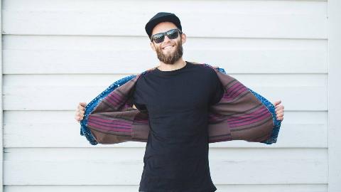 A man standing outside in sunglasses, a snapback, plain black tshirt, holding open his denim jacket as he smiles.