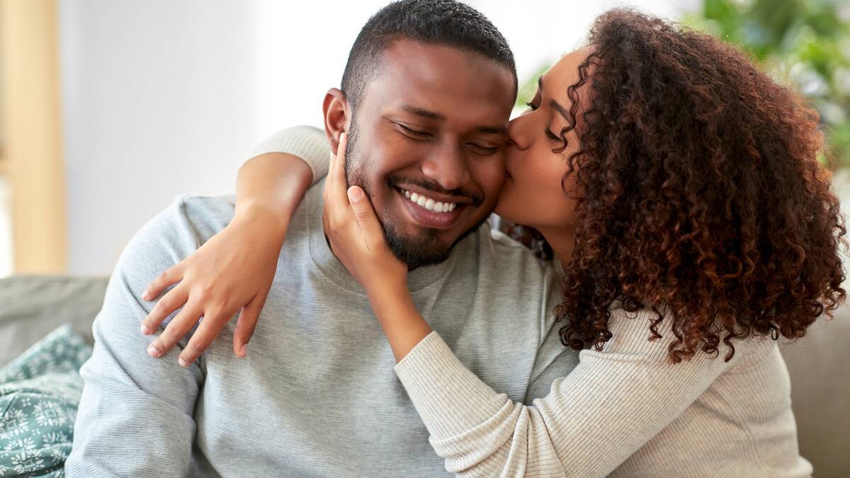 A couple sitting on a couch, the woman with her arms around the man, kissing his cheek, and cupping his jaw as he smiles.