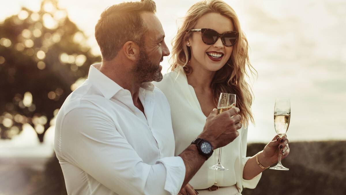 A fancy couple standing outside in the sun, each holding a glass of champagne.