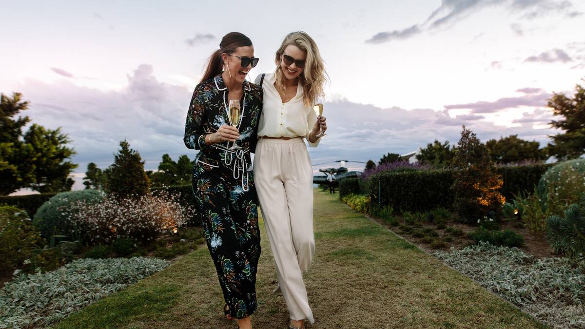 Two fancy friends walking through a garden, laughing as they chat, each holding a glass of champagne.