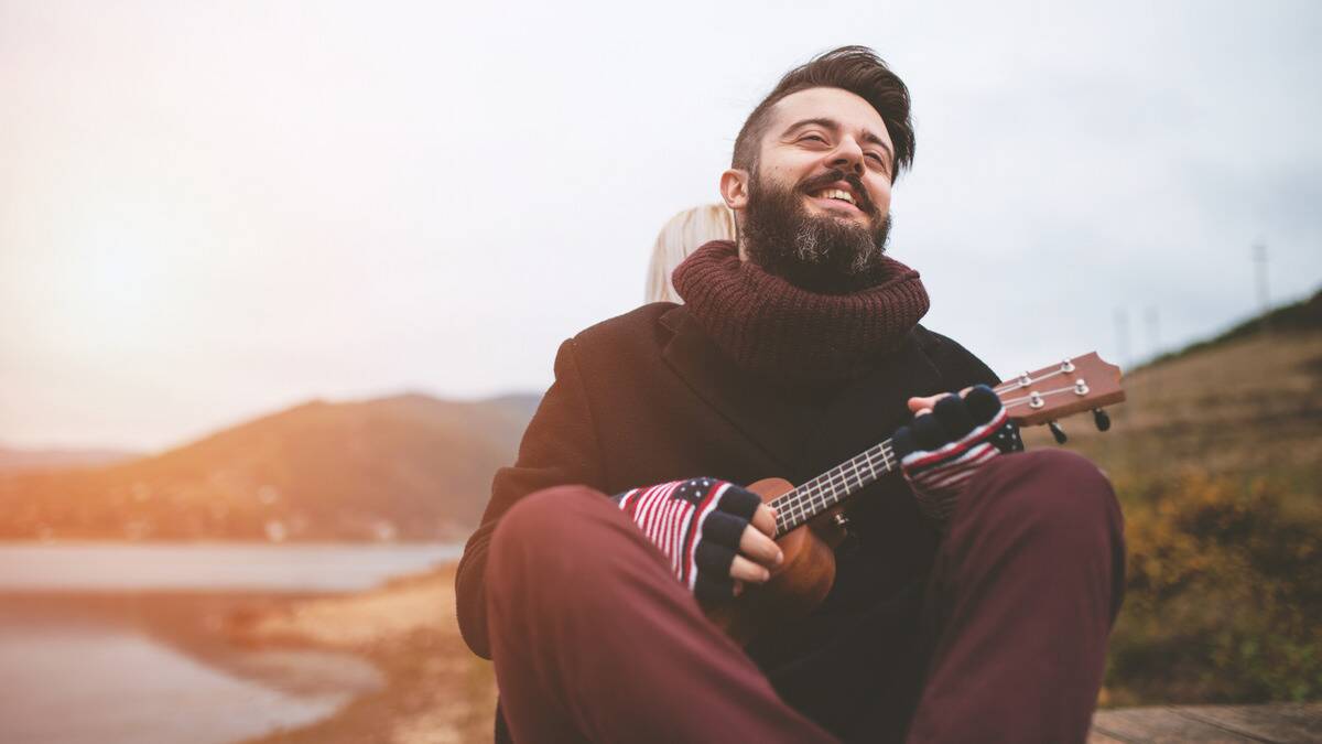 A man sitting outside, smiling as he plays a ukelele.