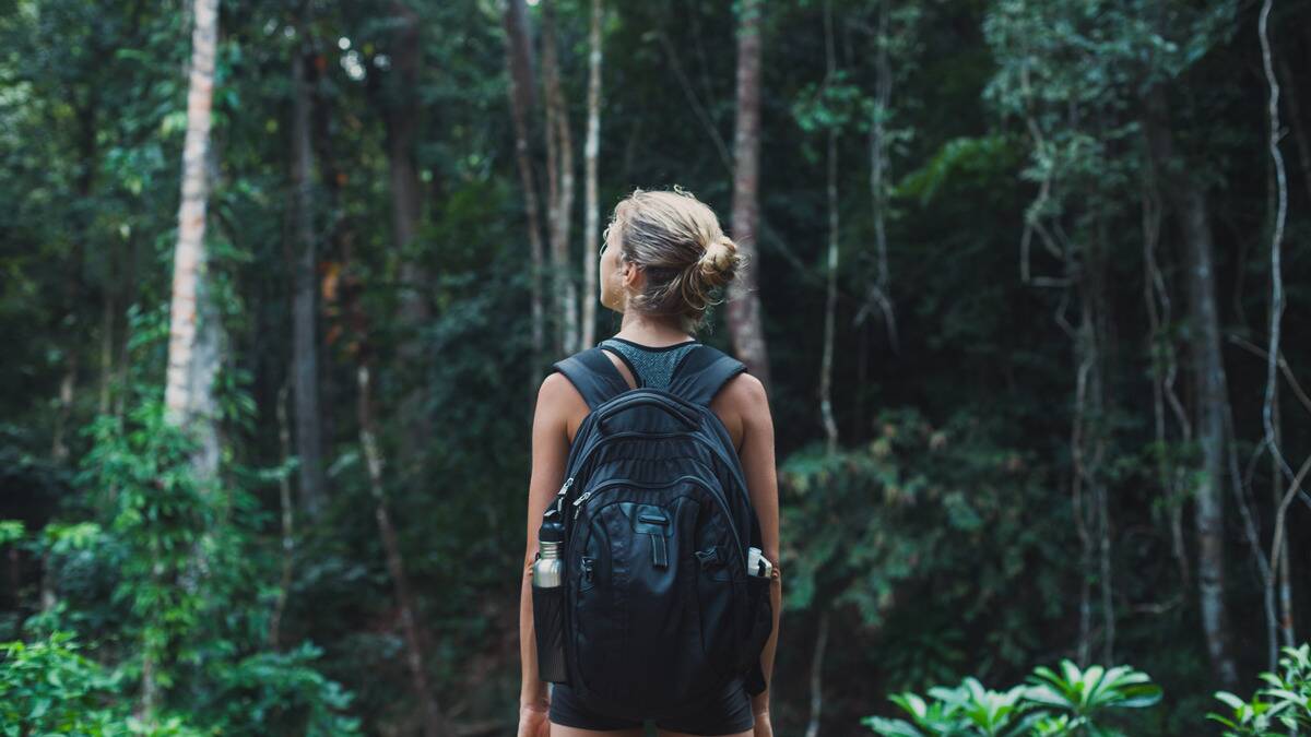 A woman standing before a thick forest, wearing a large backpack.