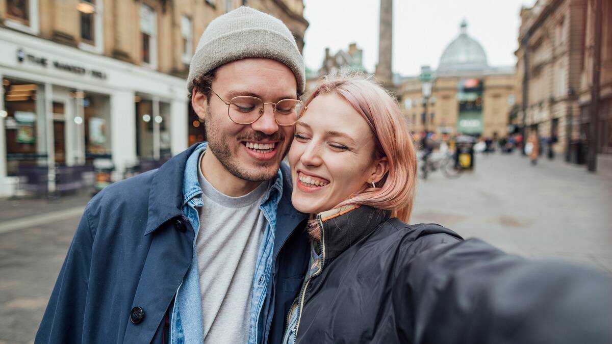 A happy young couple taking a selfie in a city street, cheeks together, both smiling with their eyes closed.