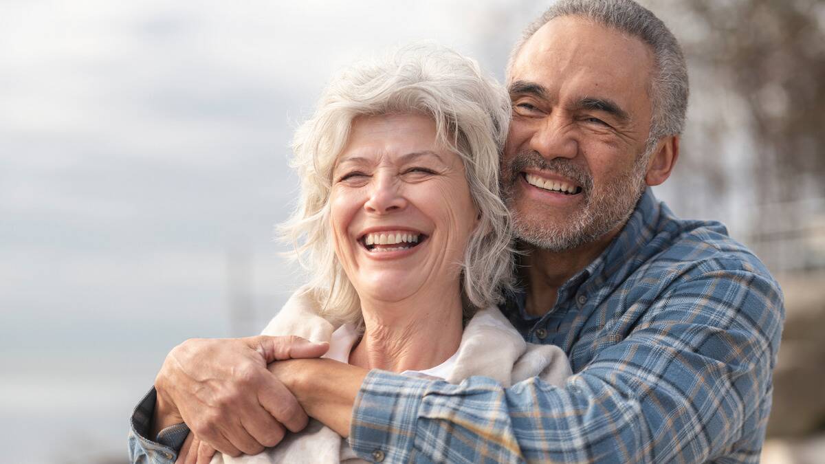 A close shot of a couple where the man is standing behind the woman, arms around her shoulders, both smiling.