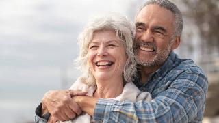 A close shot of a couple where the man is standing behind the woman, arms around her shoulders, both smiling.