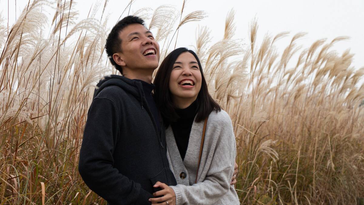 A couple standing outside by some reed grass, side-hugging and smiling as they look at something overhead.