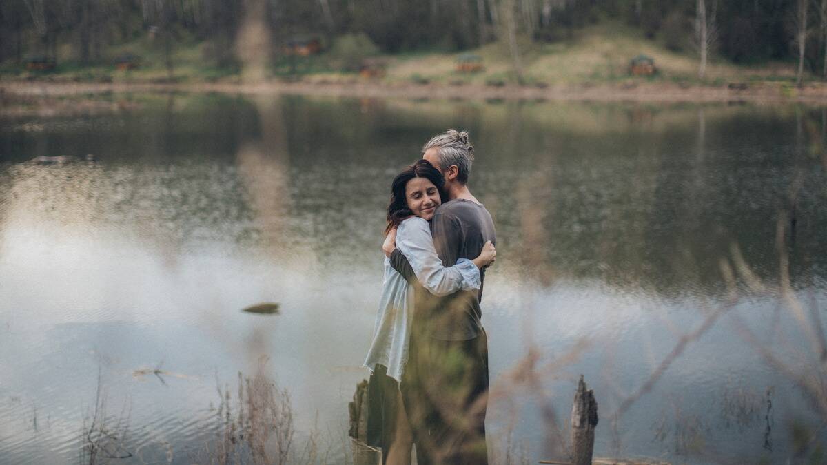 A couple hugging by the edge of the water on a lake, shot through tall grass.