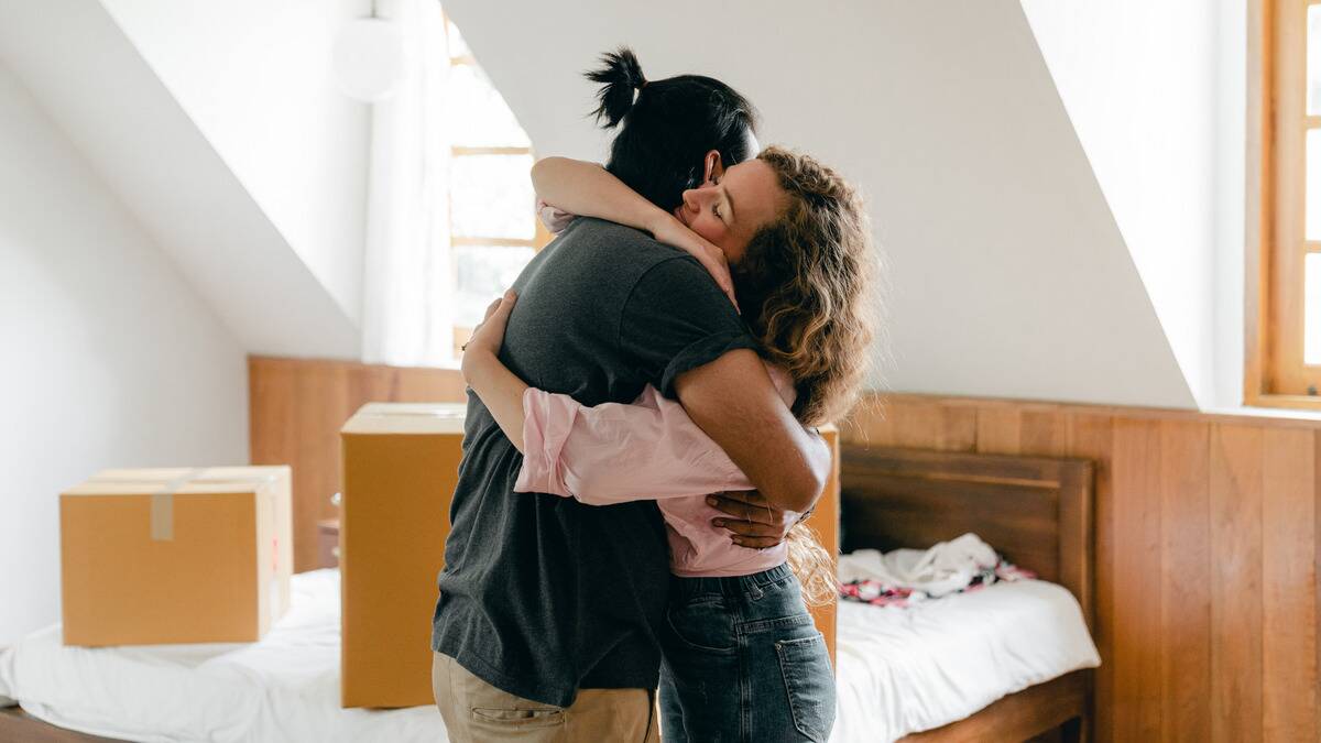 A couple hugging tightly in the bedroom of their new home, moving boxes seen in the background.