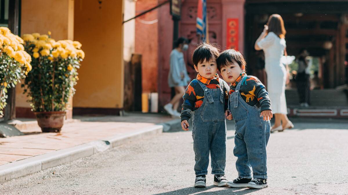 A pair of young twins in matching outfits standing in the street and looking curiously at the camera.