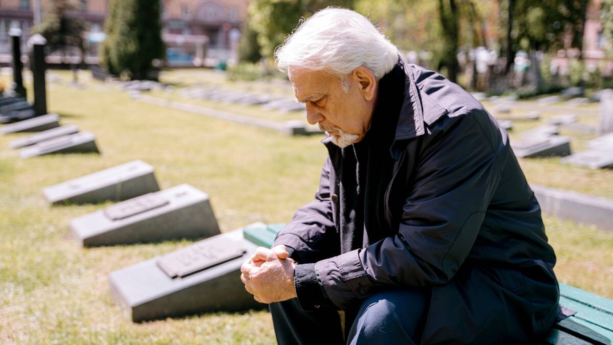 A man sitting on a bench at a cemetery, elbows on his knees, leaning forward.