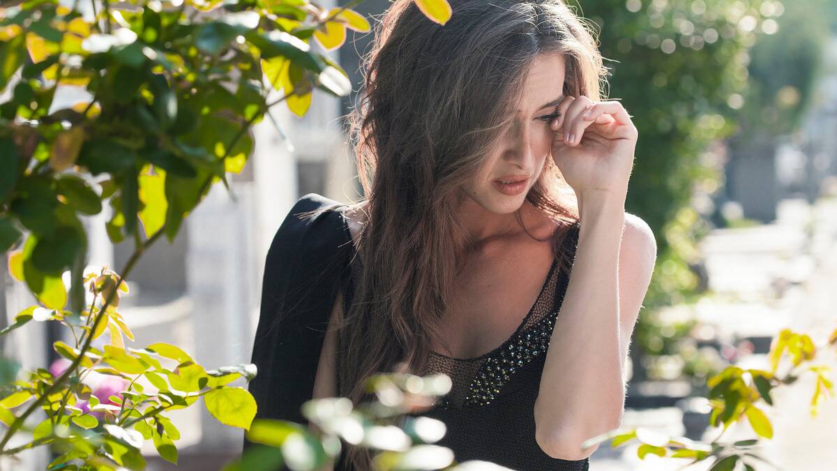 A woman crying, one hand wiping a tear away, photographed through bush branches.