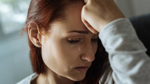 A closeup shot of a woman with a hand on her forehead, a tear on her cheek.