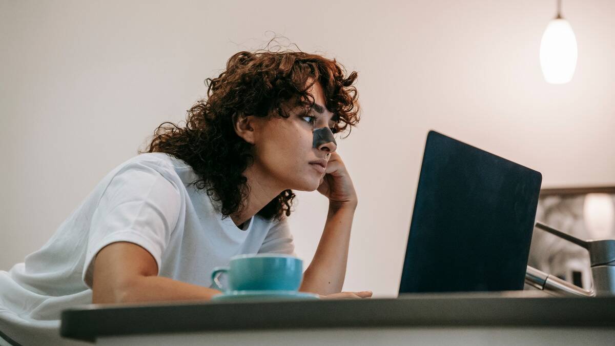 A young woman sitting at her desk, leaning forward looking at her laptop, a mug next to her and a pore strip on her nose.