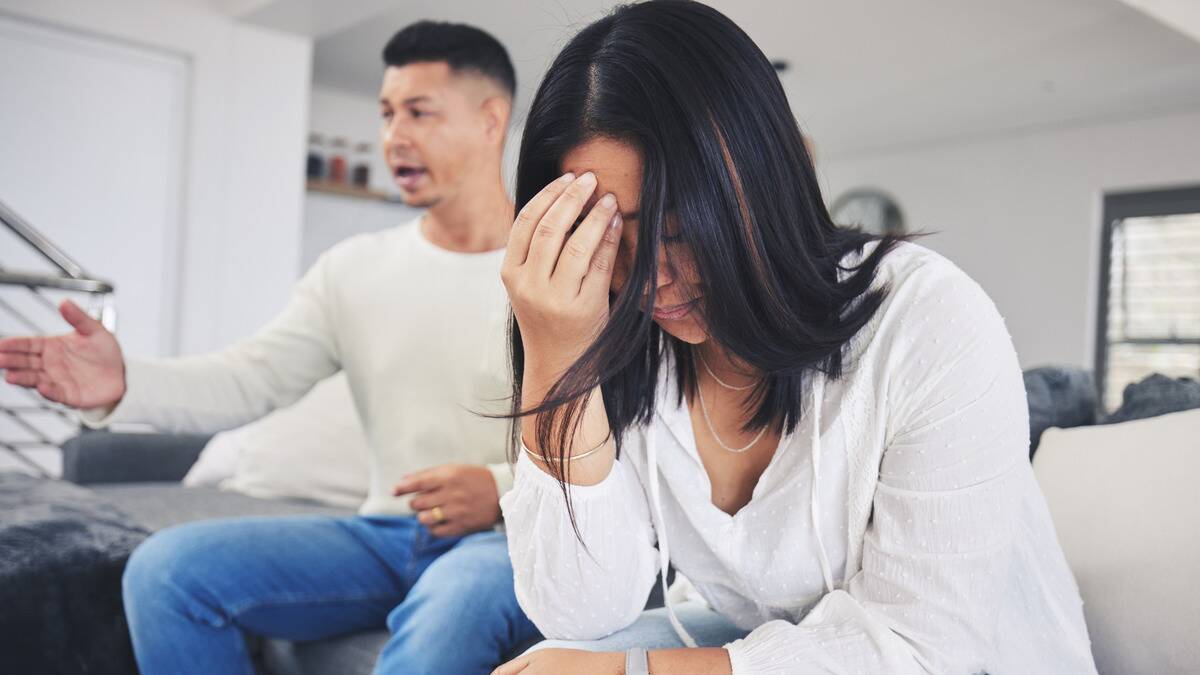 A woman sitting on her couch next to her husband, a hand on her forehead as her husband speaks critically.