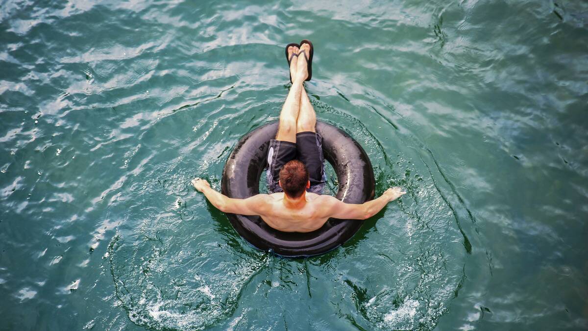 An aerial shot of a man sitting in an inner tube in swim trunks and flip flops, the water moving around him.