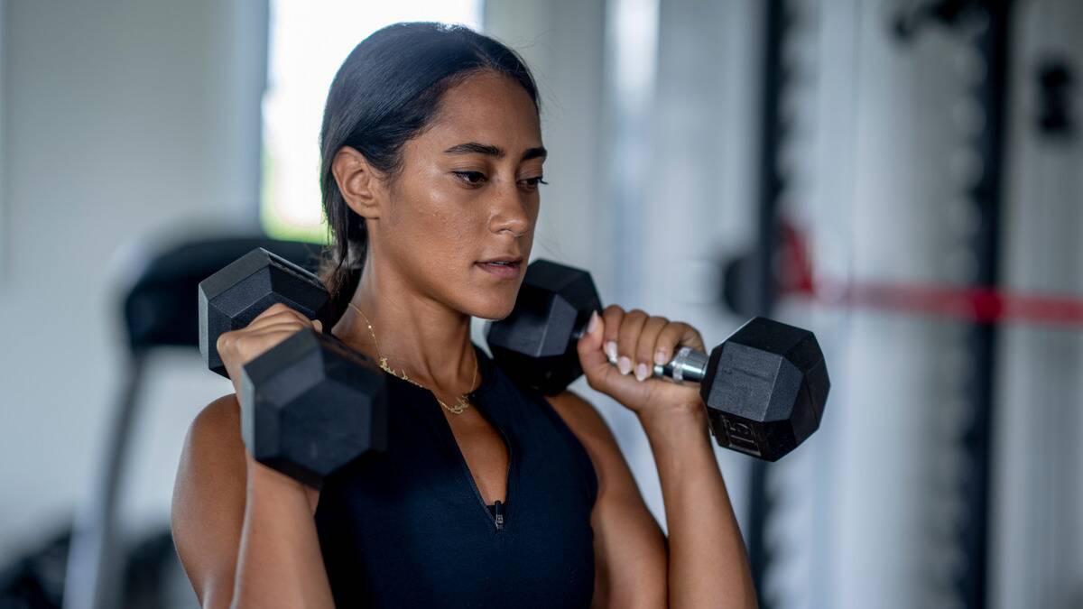 A woman looking very focused as she holds two dumbbells up at her shoulders.