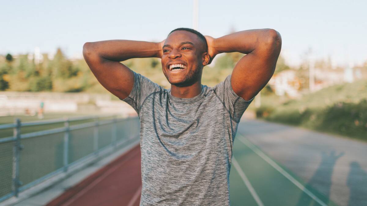 A man standing on a running track, hands behind his head, smiling confidently.