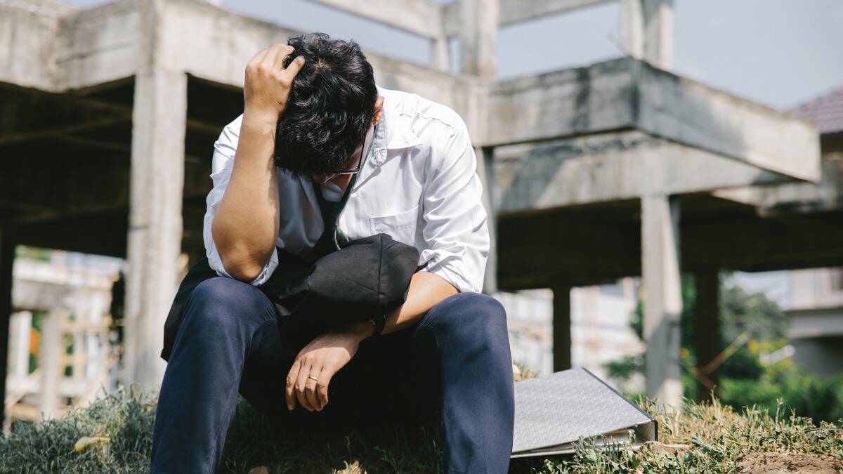 A man sitting outside with his elbows on his knees, head bowed, one hand in his hair.