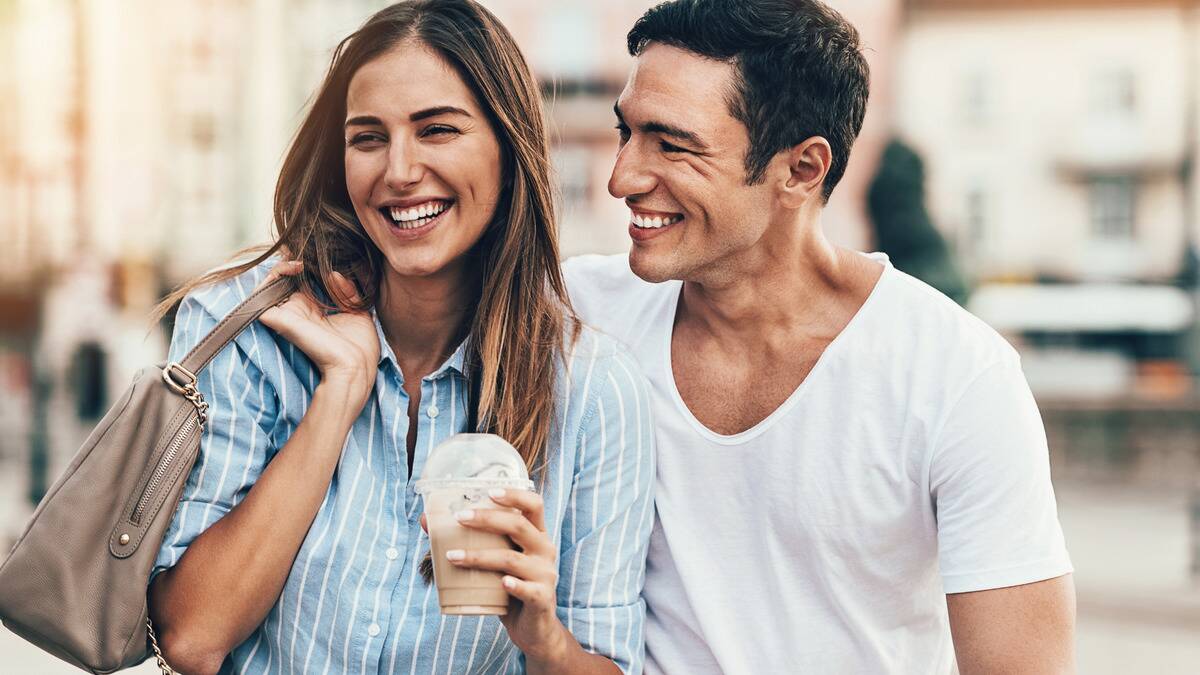 A couple out on a date, both laughing as they walk and talk, the woman holding an iced coffee.