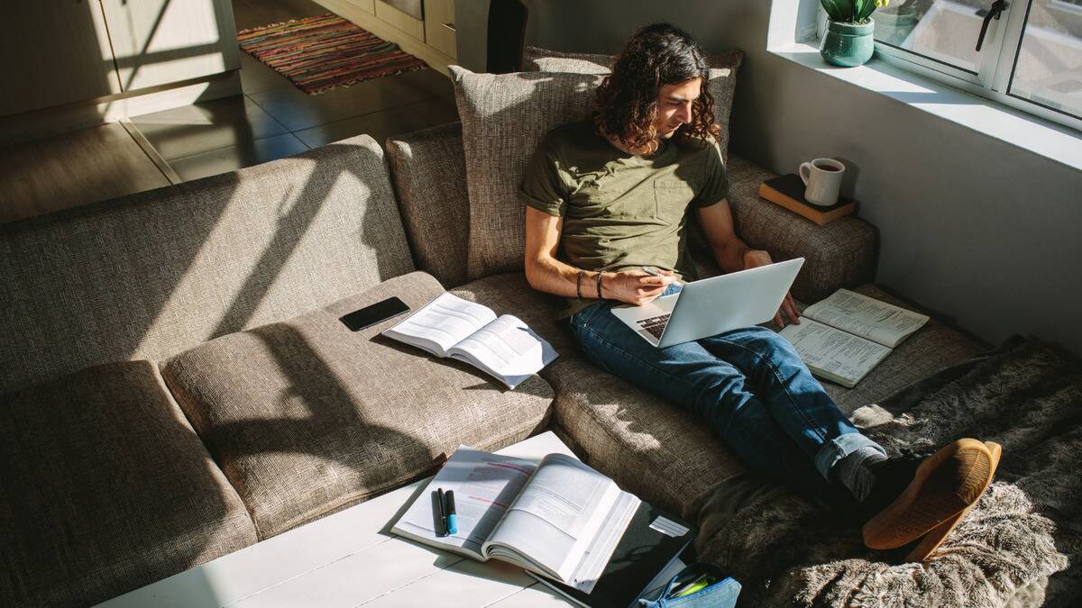 A man sat on his couch with a laptop and books scattered around him, studying something intensely.