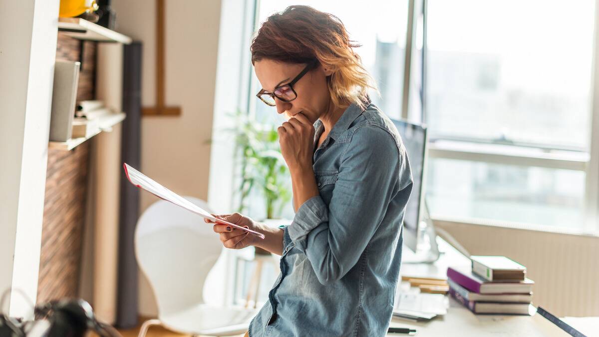 A woman standing and leaning against her desk, reading a document carefully.