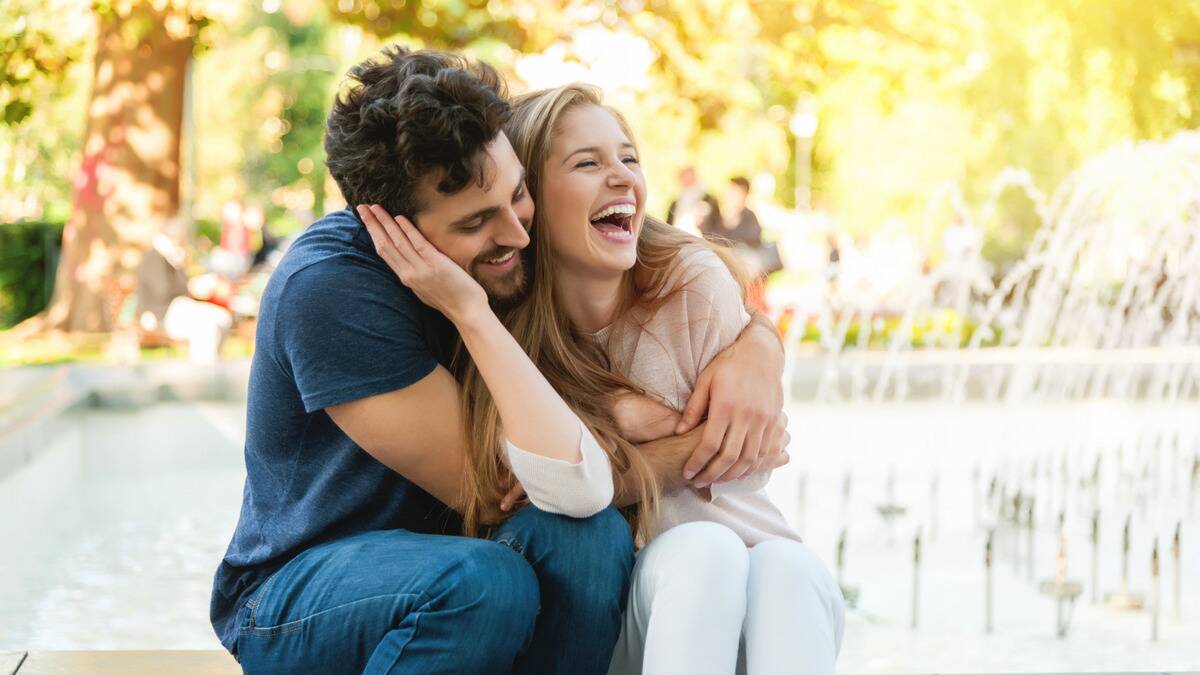 A happy couple sitting by a fountain outside, both smiling as they hold each other.