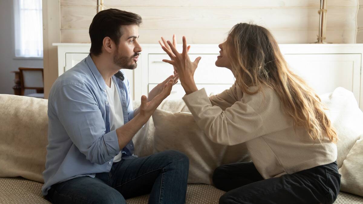 A couple sitting on a couch, facing one another and yelling.