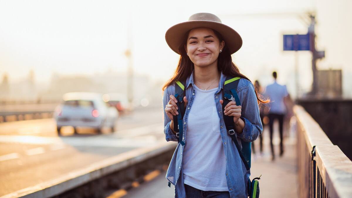 A woman walking down a city street, hands on her backpack straps, smiling at the camera.