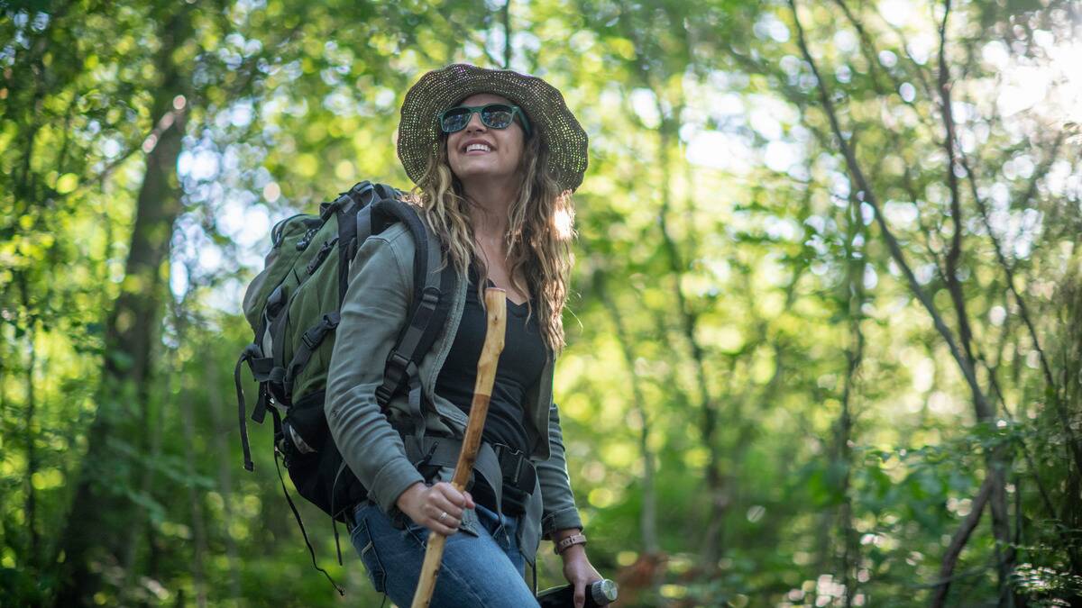 A woman hiking in the woods, using a walking stick, smiling as she looks up at the sky.