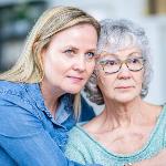 A mother and daughter sitting side by side, the daughter with her arms around her mom, but both looking forward rather seriously.