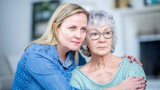 A mother and daughter sitting side by side, the daughter with her arms around her mom, but both looking forward rather seriously.