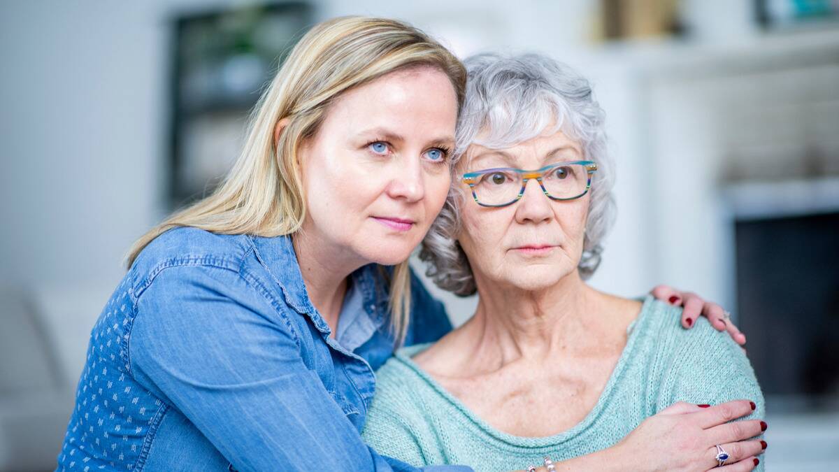 A mother and daughter sitting side by side, the daughter with her arms around her mom, but both looking forward rather seriously.