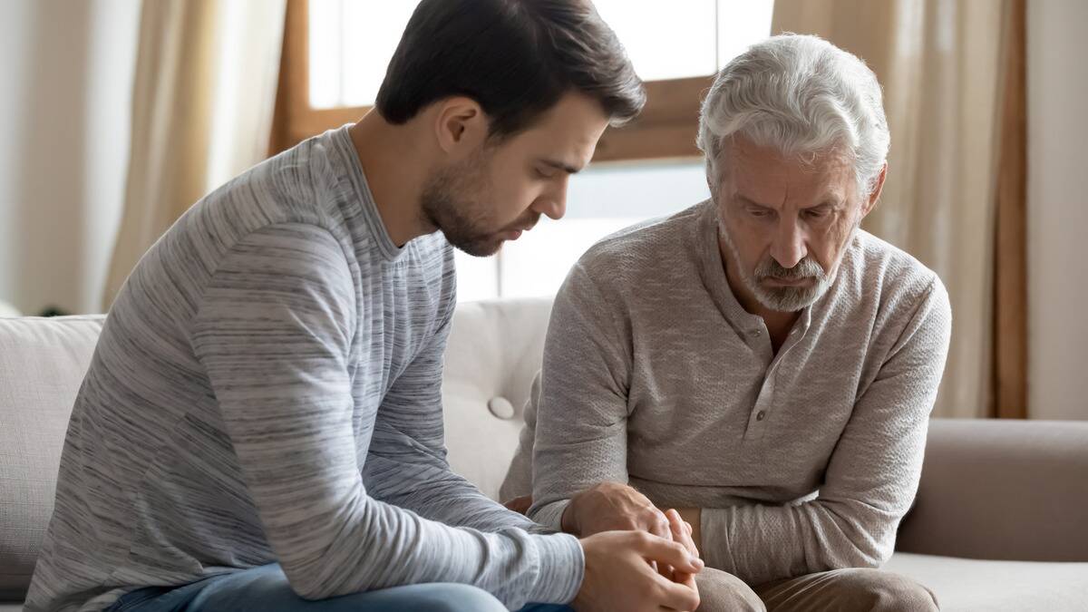 A father and son sitting side by side on a couch, both looking down in a sad fashion.