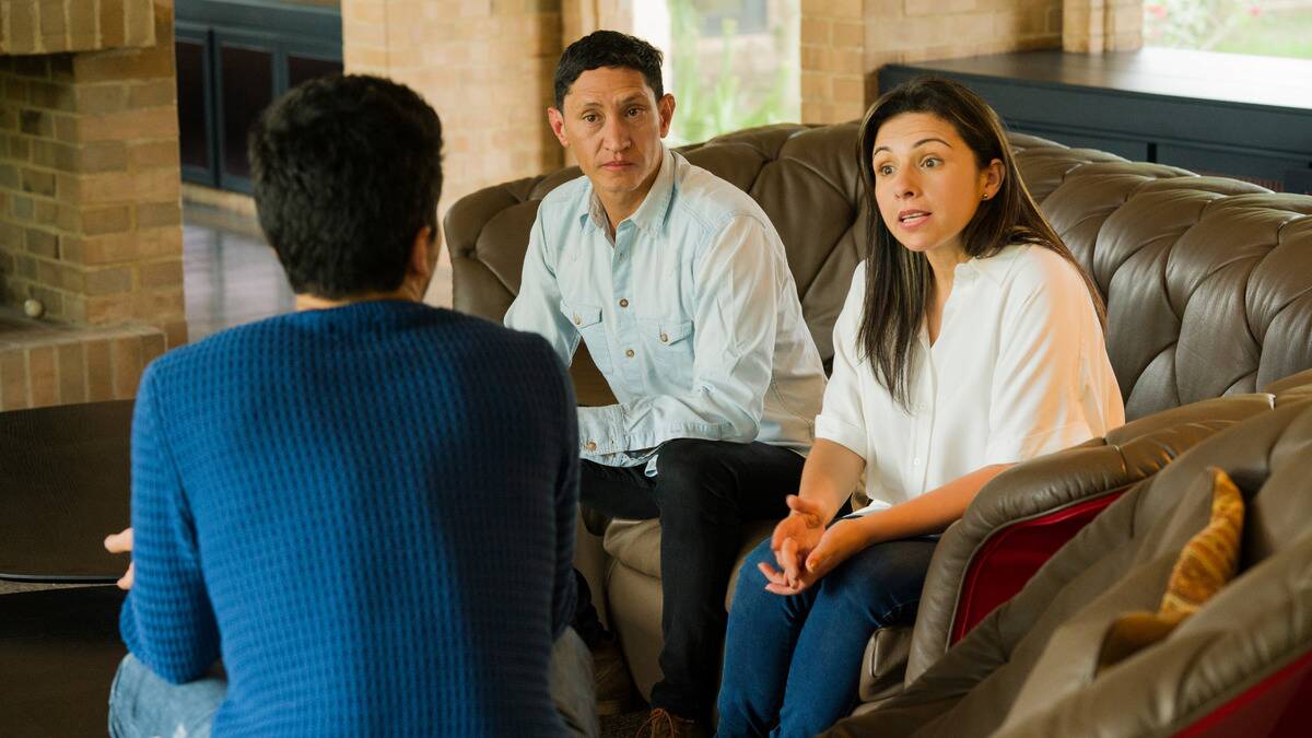 A man sitting in a chair perpendicular to his parents, telling them something. His mother looks shocked negatively, while his father looks stern.