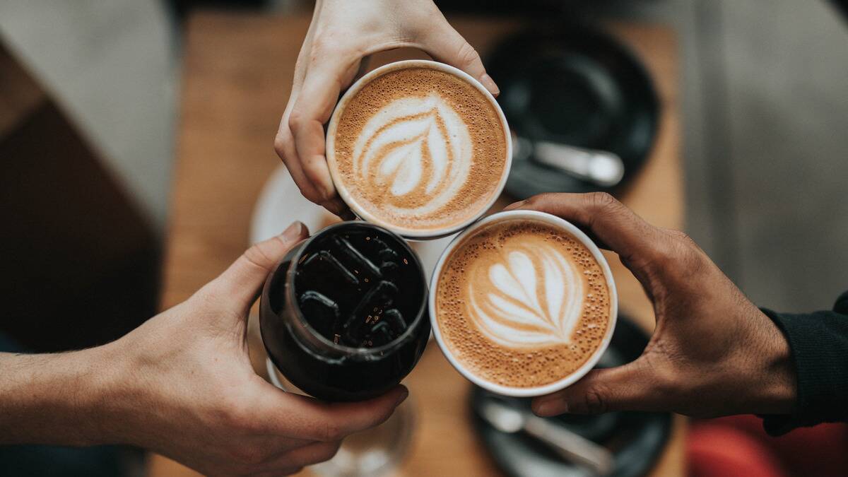 An aerial shot of three people cheersing their coffee mugs at a cafe. Two people have lattes with latte art, the third is drinking black iced coffee.