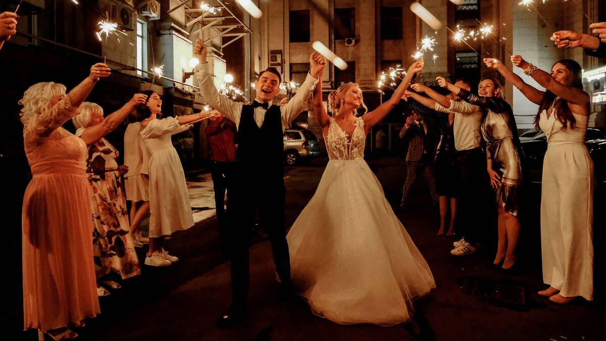 The bride and groom with their arms in the air, smiling and celebrating as their guests hold sparklers above them.