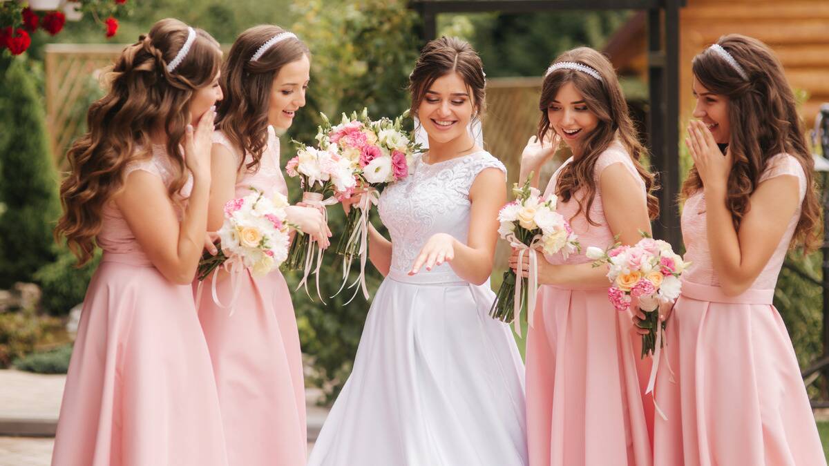 Four bridesmaids in pink dresses gathering around the bride who's showing off her wedding ring, all smiling and holding bouquets.