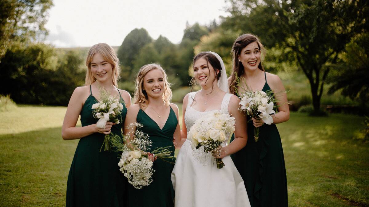 A bride standing outside with three of her bridesmaids, who are all in dark green dresses, each of them carrying a bouquet and smiling.