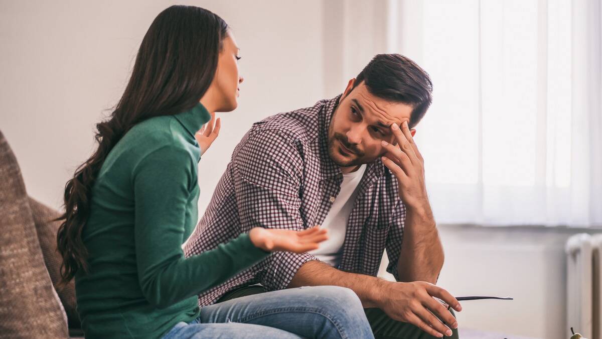 A man looking exacerbated, one hand on his temple, as his girlfriend speaks to him.