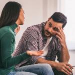 A man looking exacerbated, one hand on his temple, as his girlfriend speaks to him.