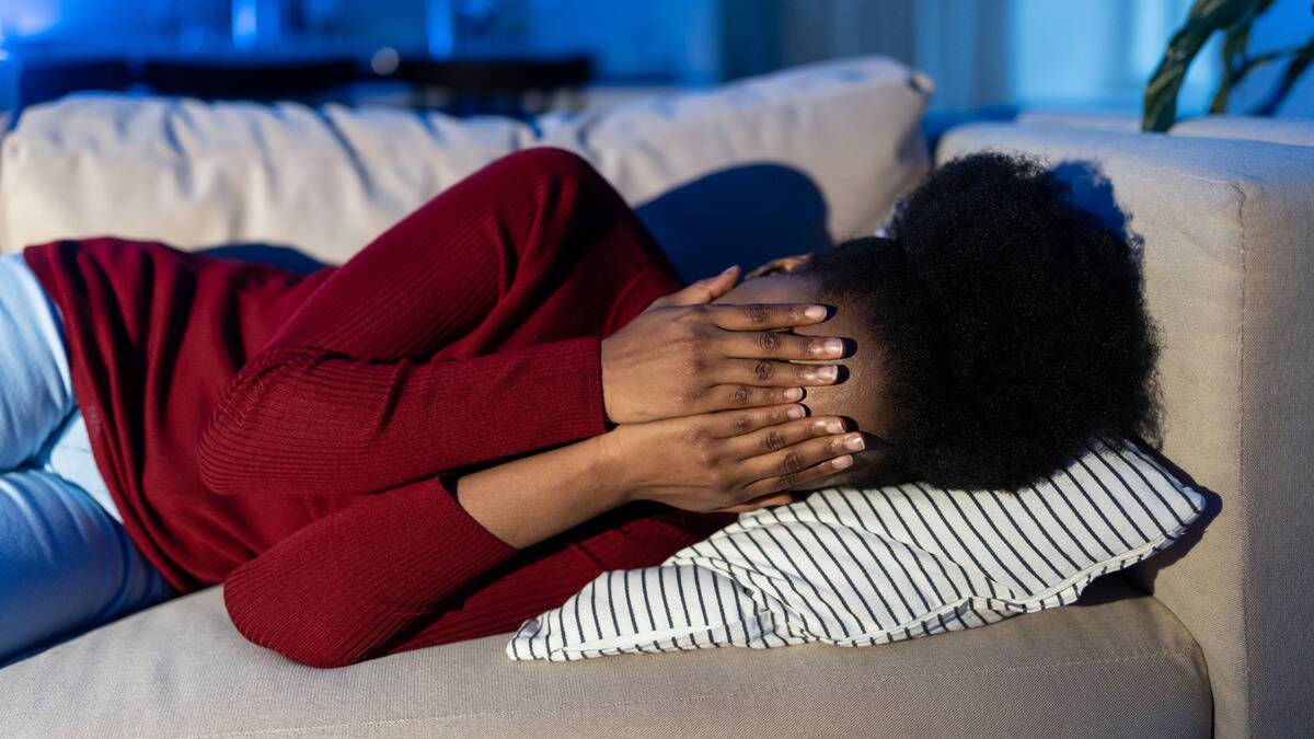 A woman laying on her couch on her side, covering her face with her hands.